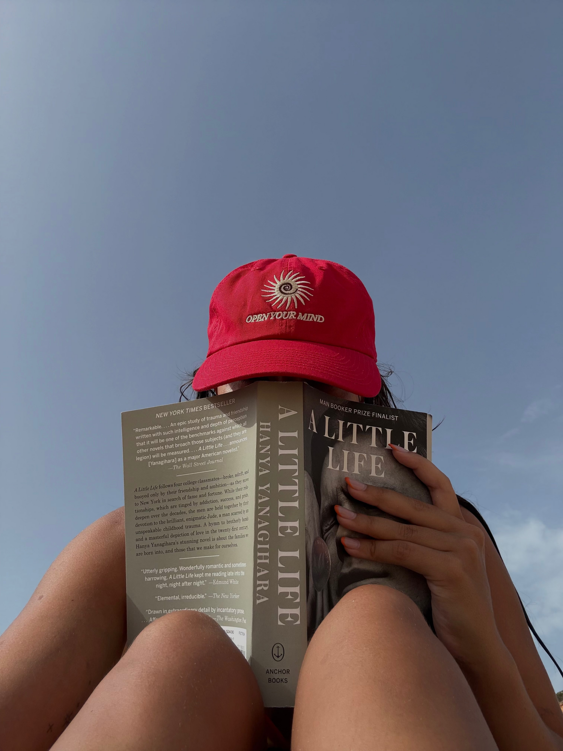 Person reading 'A Little Life' with a red cap on a clear sky background